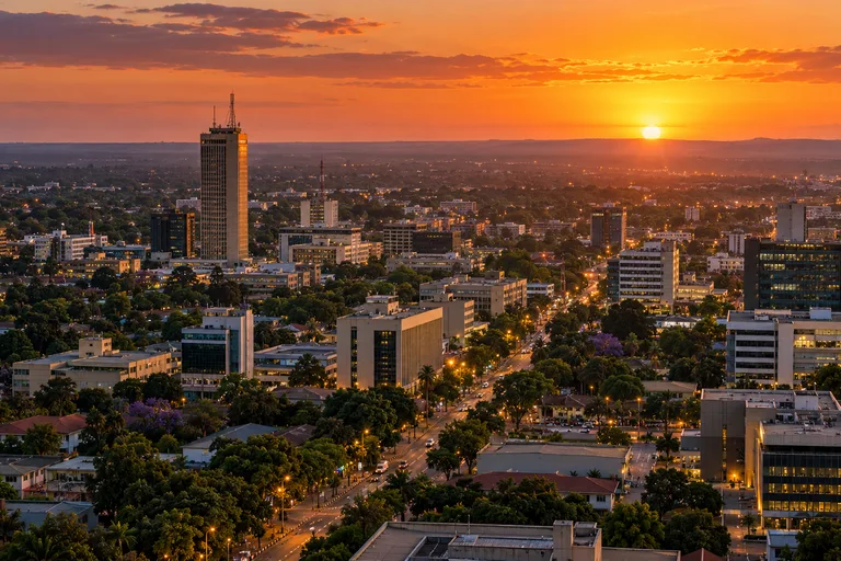 AI-generated illustration of Lusaka’s skyline at sunset, with office buildings and tree-lined streets beneath an orange sky.